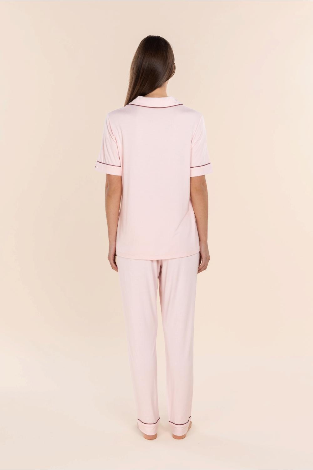 A woman viewed from the back wearing a light pink short-sleeve polo pajama set with thin dark red piping on the collar and sleeve cuffs, paired with matching light pink pajama pants, against a solid pale beige background.