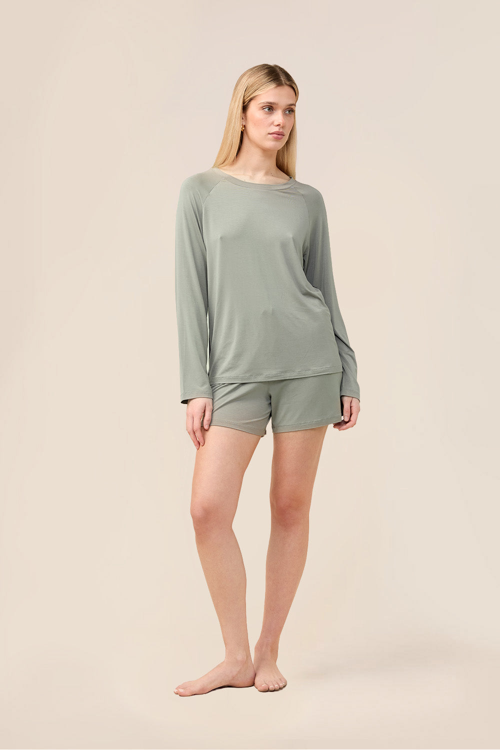 A woman modeling a matching sage green long-sleeve top and shorts pajama set against a solid light beige studio background.