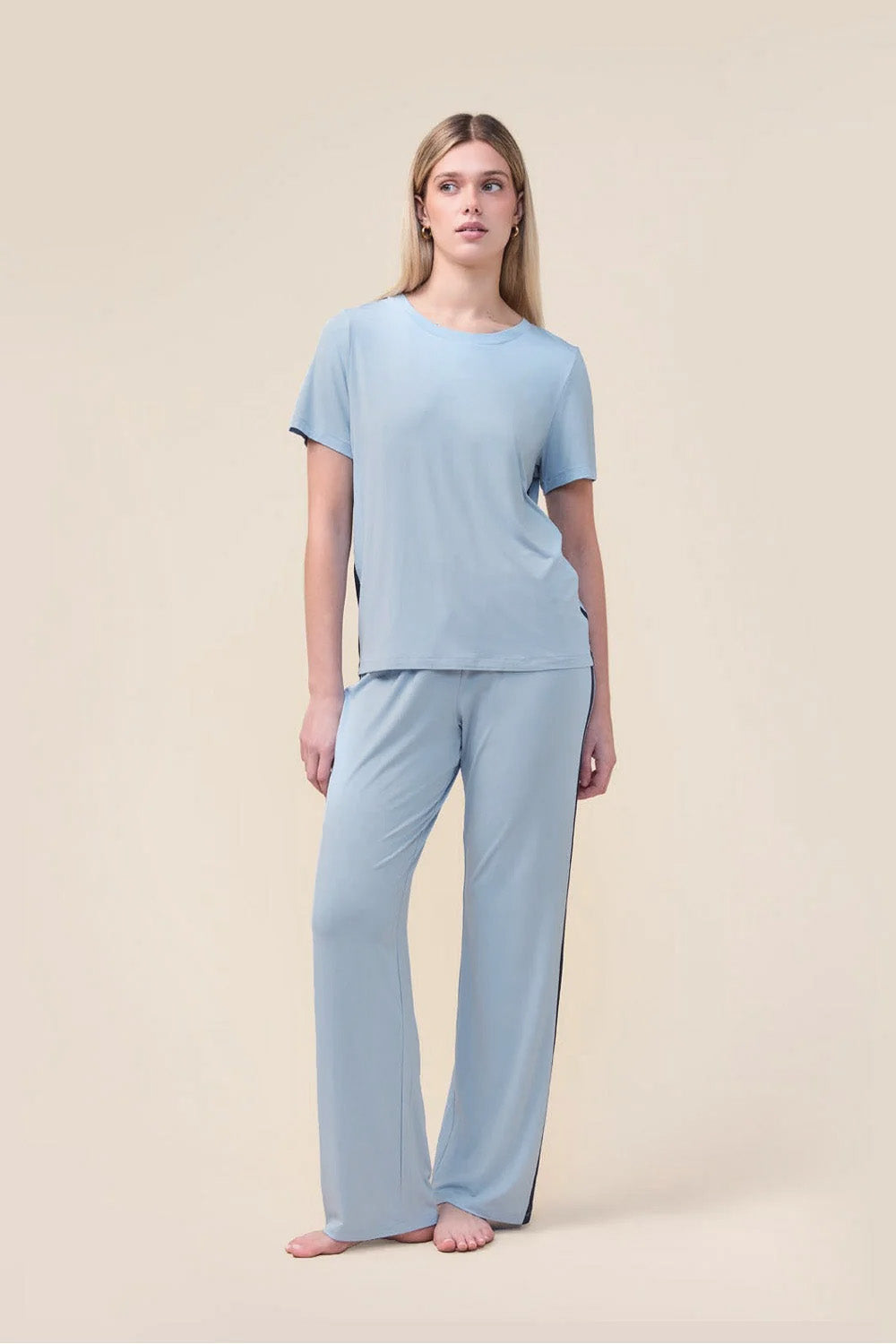 A woman modeling a light blue two-piece pajama set consisting of a short-sleeve crewneck top and matching wide-leg pants, standing against a solid beige studio background.