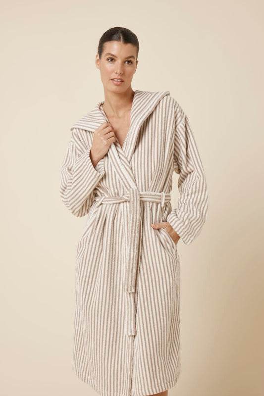 Slight close up view of woman wearing a striped adriana terry towel robe in biscuit colour standing against a plain background with hand in pocket and on upper neckline