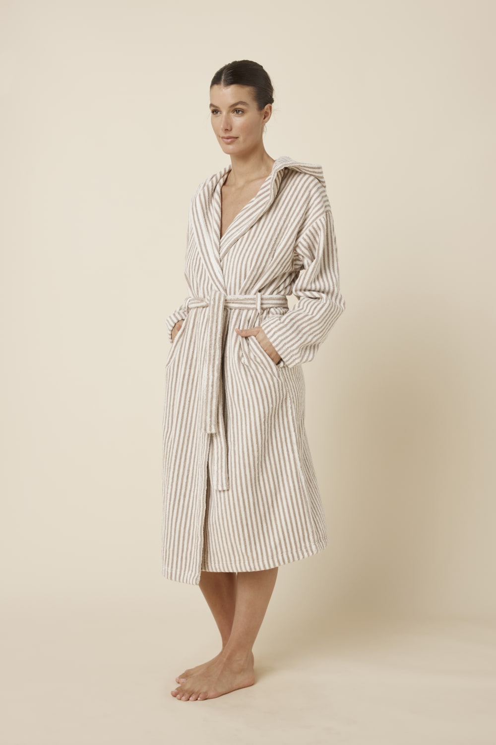 Woman wearing a striped adriana terry towel robe in biscuit colour standing against a plain background from a side on view