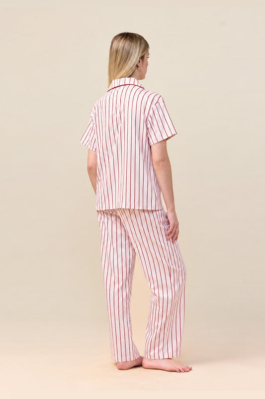 A woman stands barefoot from the back wearing a short-sleeve collared pajama top and matching pajama pants with vertical red and white stripes, against a plain beige studio background.