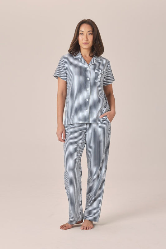 A woman modeling a navy and white striped pyjama set with a short-sleeve button-up top and long pants, standing against a solid beige studio background.