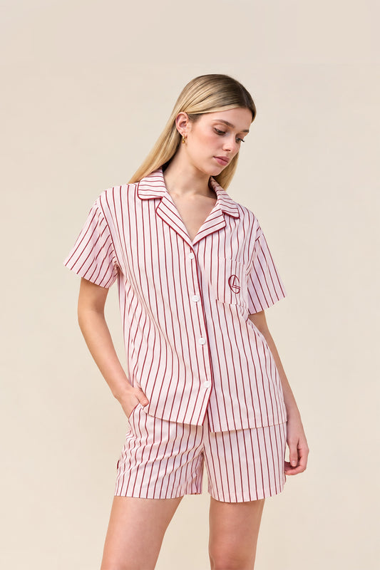 A woman modeling a matching short-sleeve button-up pajama shirt and shorts set with vertical red stripes on a warm off-white background, photographed against a solid beige studio backdrop.