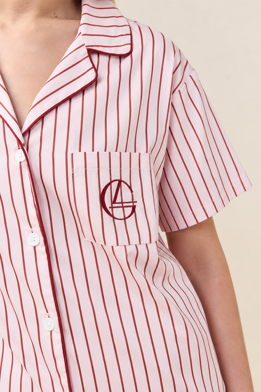 A close-up of a person wearing a short-sleeve button-front shirt with thin red vertical pinstripes on a white base. The shirt has a chest pocket with a matching red embroidered monogram logo and is photographed against a plain light beige background.