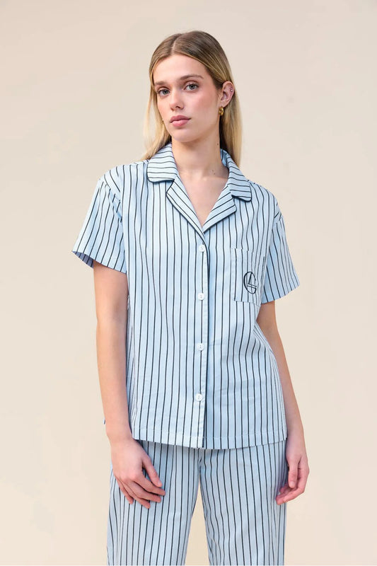 A woman modeling a light blue and navy striped short-sleeve pajama shirt and matching pants against a solid beige studio background.