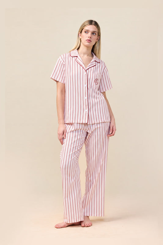 A woman modeling a two-piece pajama set with short-sleeve button-up top and matching pants, featuring vertical pink and white stripes on a warm beige studio background.