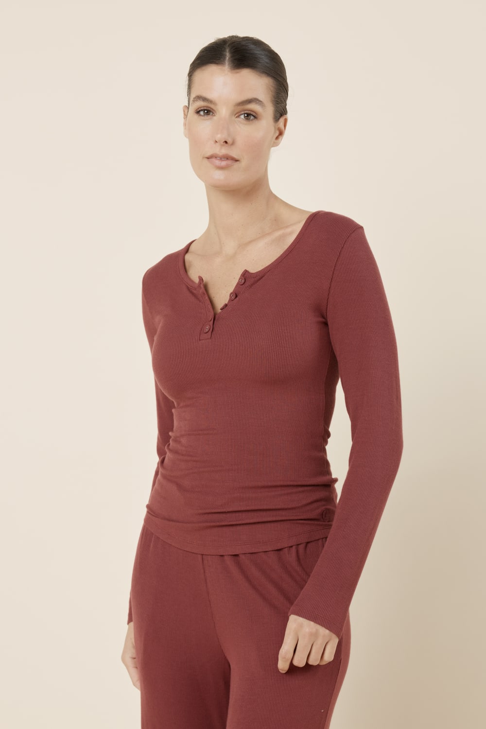 Woman wearing Lille Ribbed Long Sleeve Henley Top Burgundy standing full length, showing the slim silhouette, curved hem and classic henley button front.