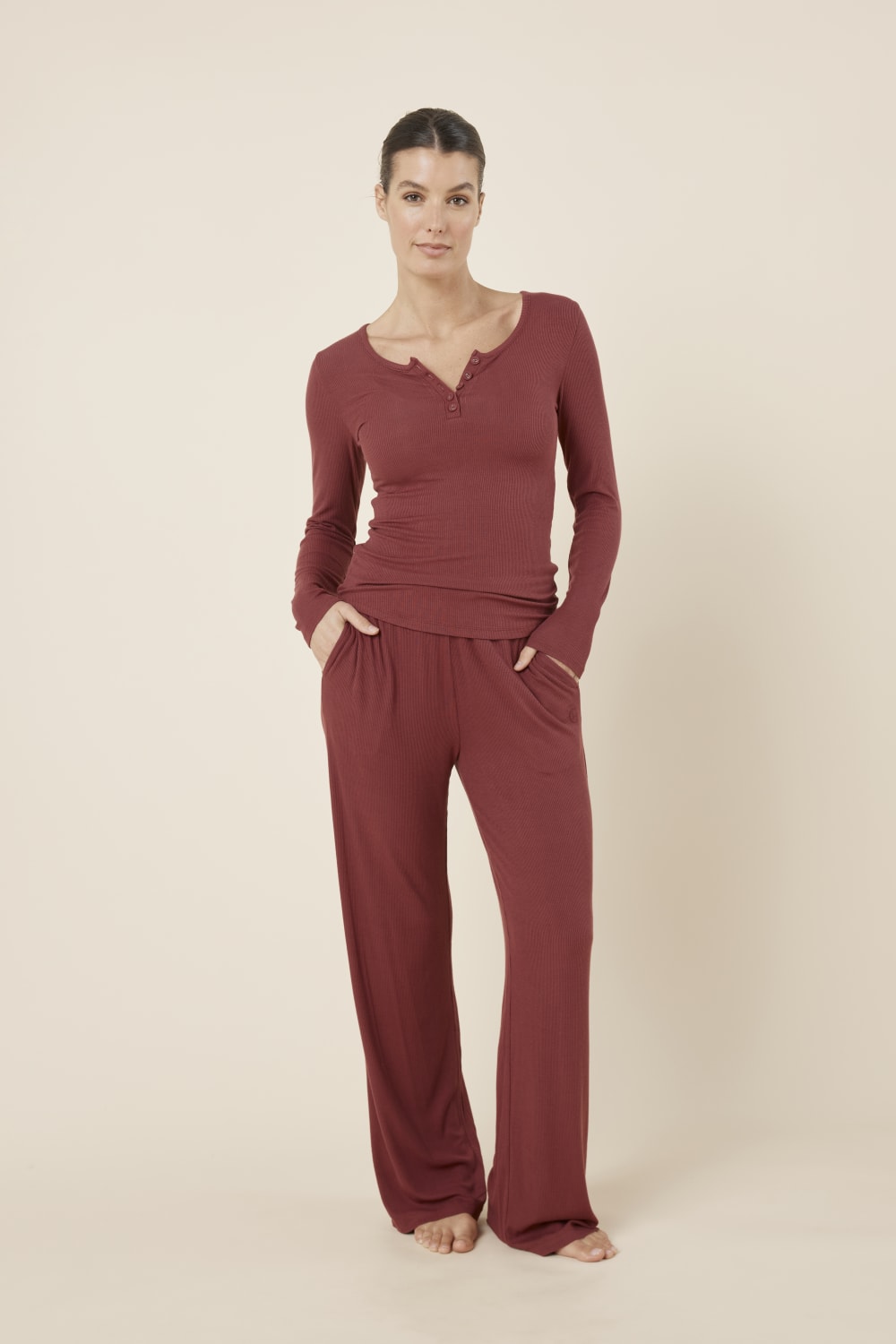 Woman wearing Lille Ribbed Long Sleeve Henley Top Burgundy standing in matching burgundy bottoms, showing the coordinated lounge set and fitted ribbed fabric.