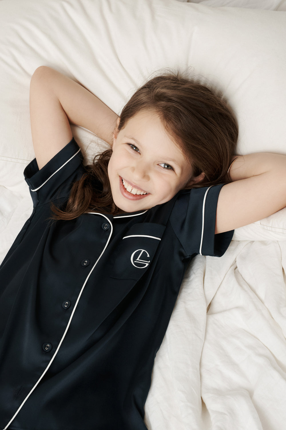 Smiling girl in navy Gingerlilly Sleepwear pajamas lying on a white bed, natural fabrics