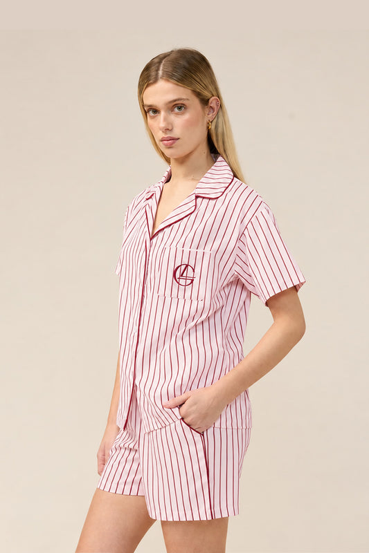 A woman modeling a short-sleeve pajama set with pink and white vertical stripes, featuring a button-down shirt and matching shorts with a red embroidered monogram on the chest pocket.