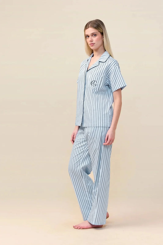A woman modeling a two-piece pajama set with light blue and white vertical stripes, standing against a solid beige studio background.