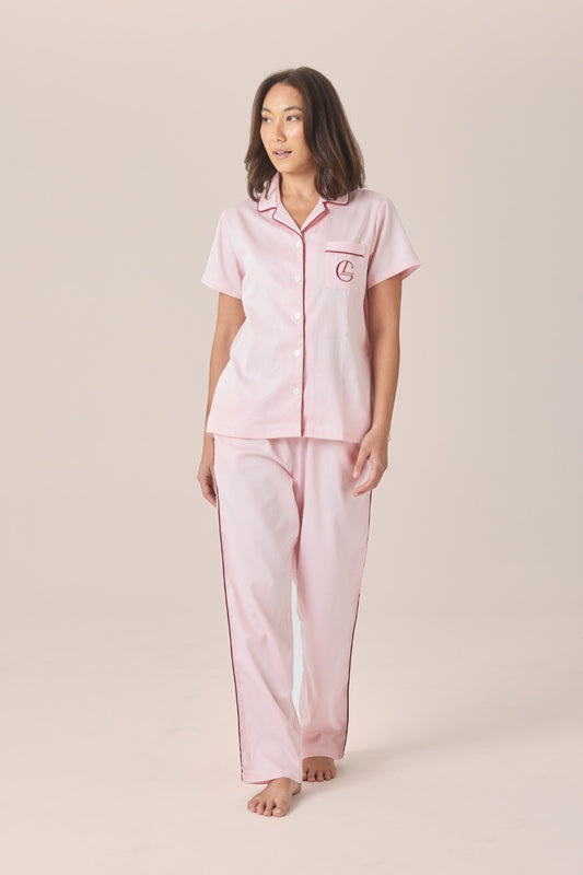 A woman modeling a matching pale pink pyjama set with a short-sleeve button-front top and long pants, featuring an embroidered logo pocket, against a solid beige studio background.