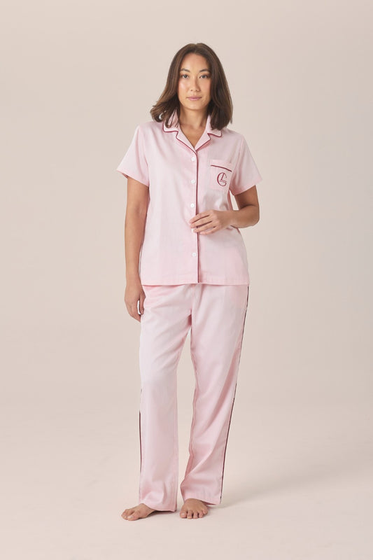 A woman modeling a pale pink pyjama set with a short-sleeve button-up top and long pants, standing against a solid beige studio background.