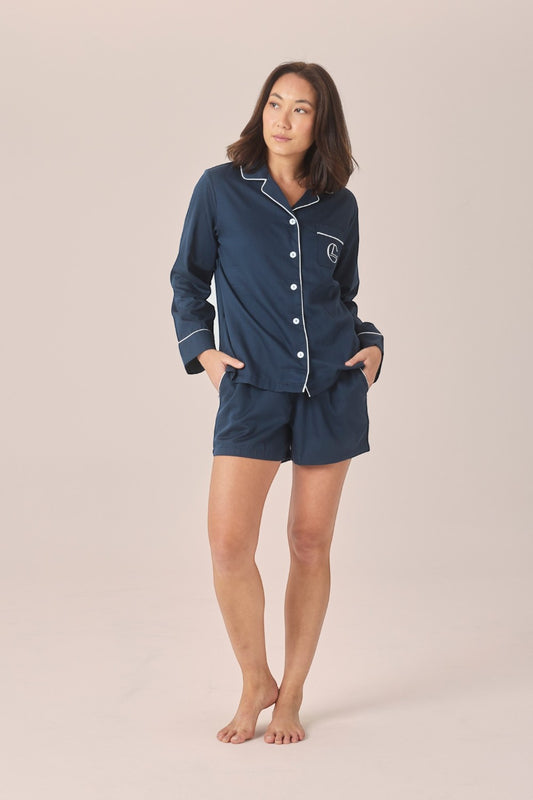 A woman modeling a navy blue pajama set with white piping details, consisting of a long-sleeve button-up shirt and matching shorts, against a solid light beige background.
