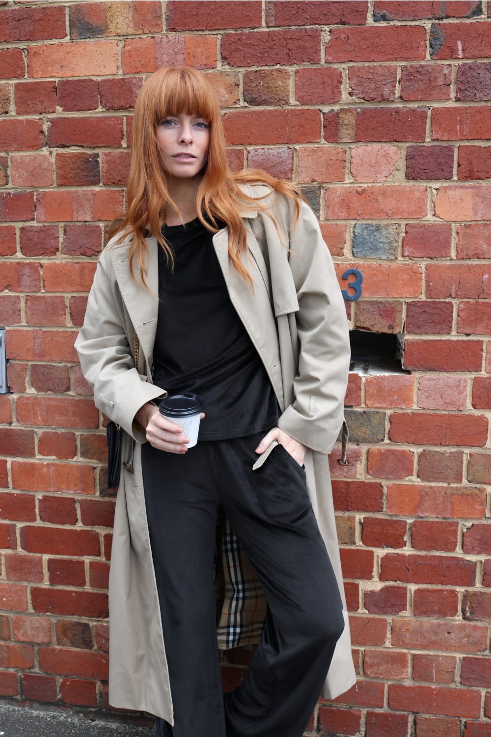 Woman with red hair in black loungewear and beige trench coat holding coffee, brick wall background, Gingerlilly Sleepwear