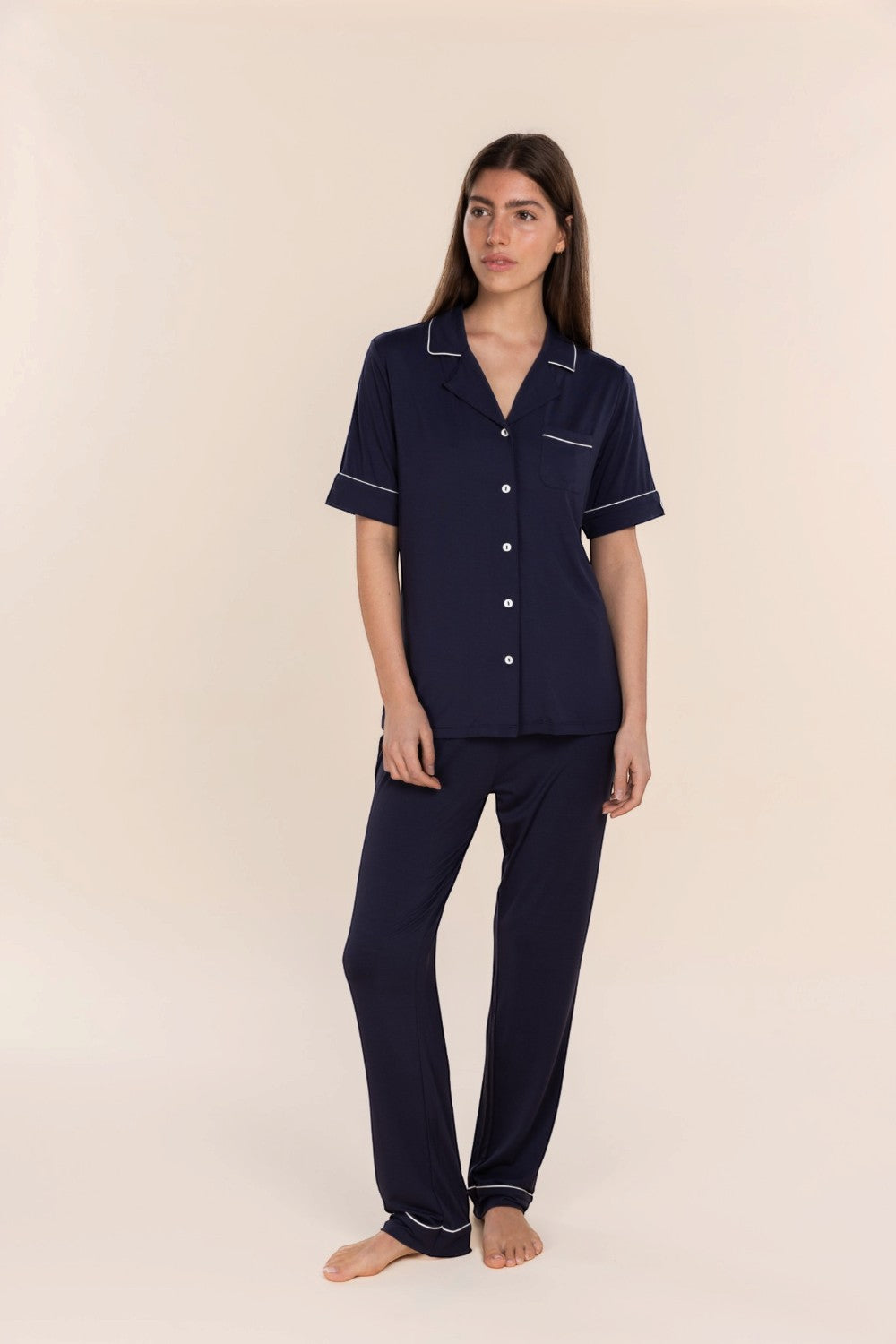 A woman modeling a navy blue two-piece pajama set with white piping details, consisting of a short-sleeve button-up shirt and matching pants, against a solid light beige background.