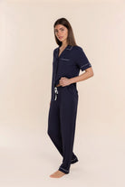 A woman modeling a navy blue short-sleeve pajama set with white piping and a white drawstring, standing against a solid light beige background.