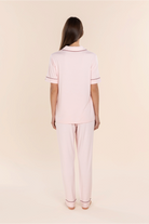A woman viewed from the back wearing a light pink short-sleeve polo pajama set with thin dark red piping on the collar and sleeve cuffs, paired with matching light pink pajama pants, against a solid pale beige background.